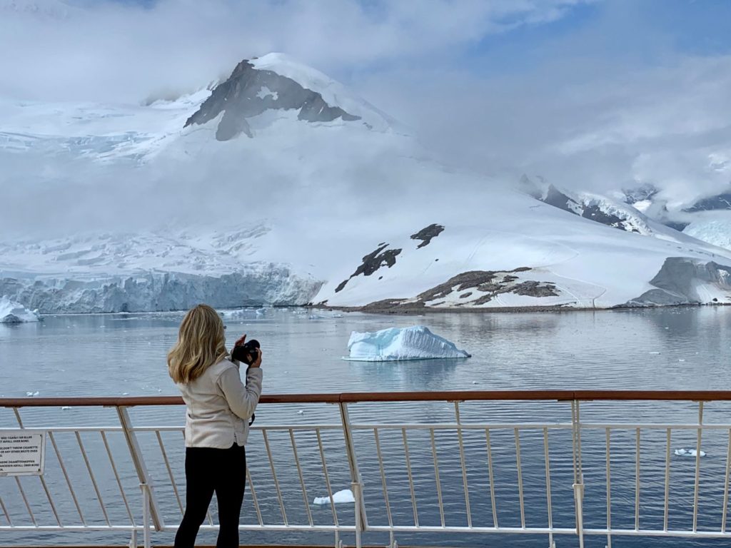 Jackie J.Reid standing on ship deck with a camera looking out at Neko Harbor, Antarctica.